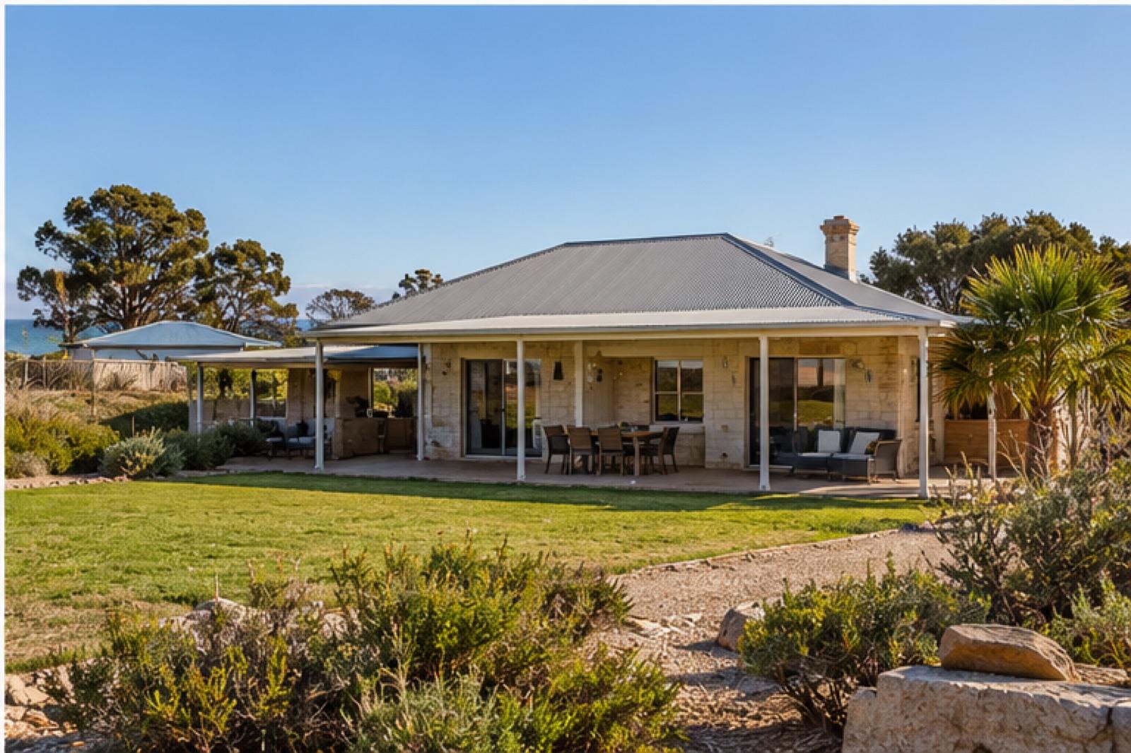 A single-storey limestone house with a deep wraparound verandah, lawn in front of it and the sea visible beyond the trees.